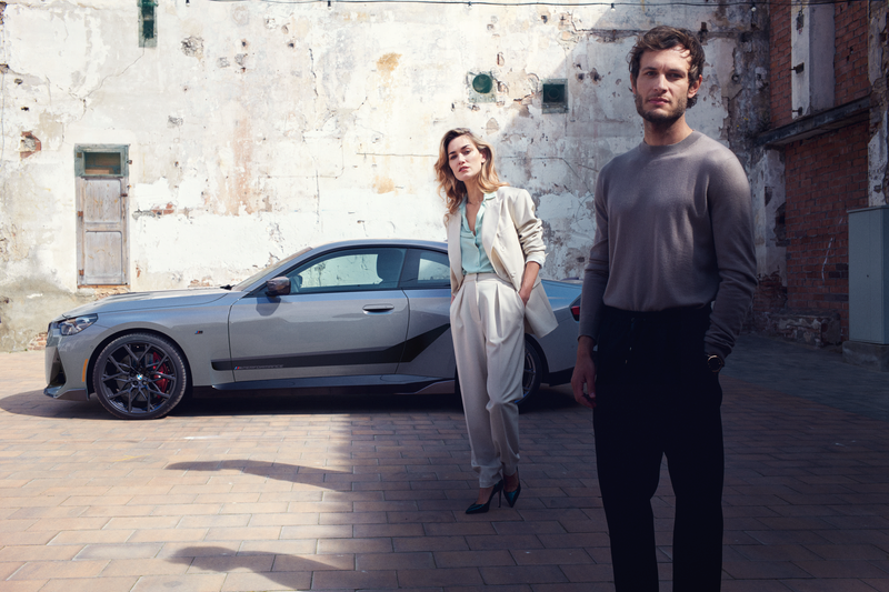 Side view of a BMW 2 Series G42 parked outside next to a building with a male and female posing in the foreground. Vehicle outfitted with M Performance Parts, including: 20-inch forged wheel Y-spoke 795 M Jet Black matte summer complete wheel set, 18-inch brake system, M Performance decals in Frozen Black, and carbon fiber front attachment, mirror cap and side skirt blade.